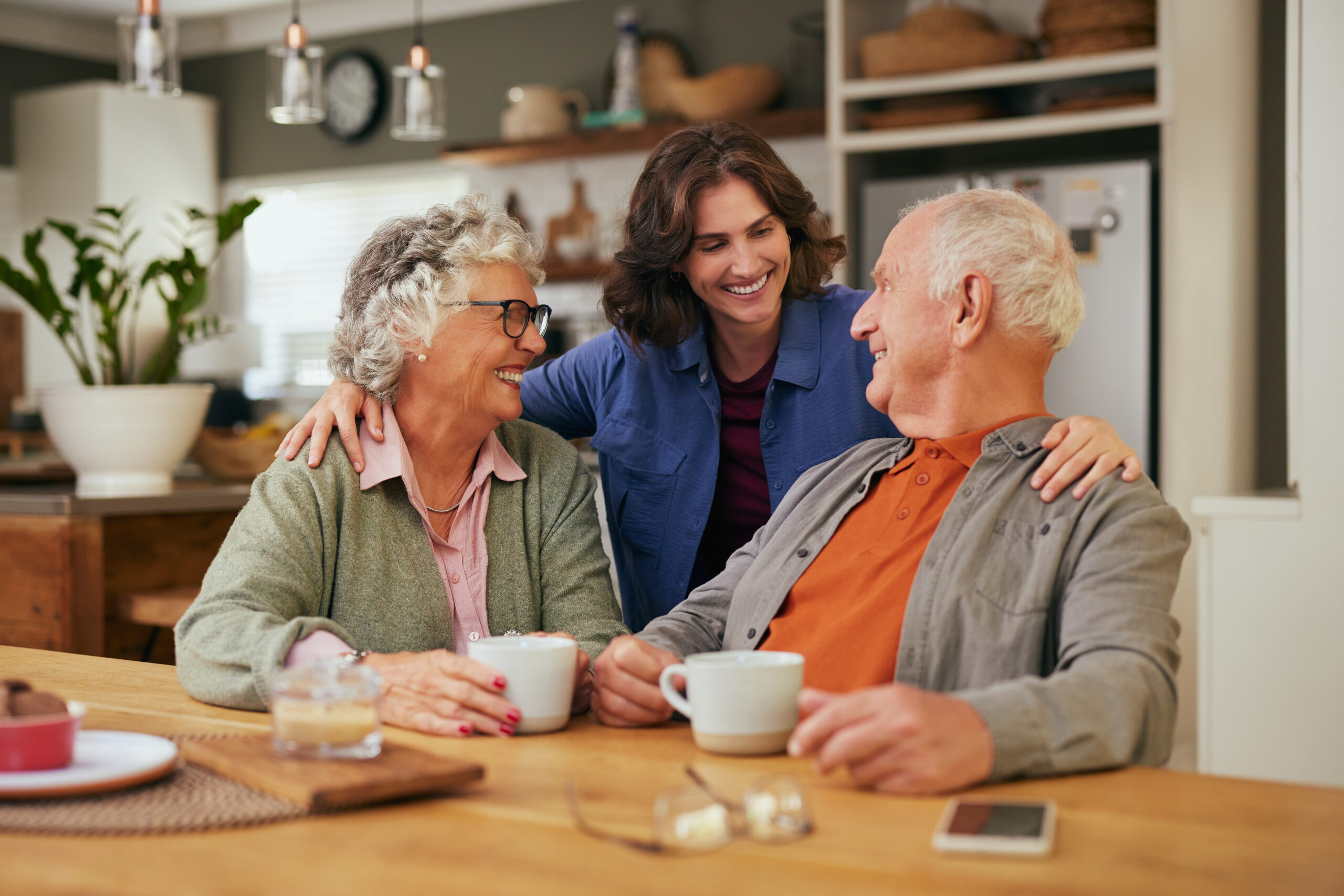 Senior father and old mother sharing tea and heartfelt conversation with daughter at home at tea time. Elderly parents and beautiful daughter bonding over tea and discussion. Warm daughter embracing from behind her joyful parents while sitting at table and having a good conversation.